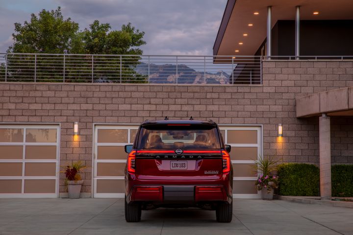 Rear view of a red 2025 Nissan Armada parked in front of the garage attached to of a modern stone mansion at dusk.
