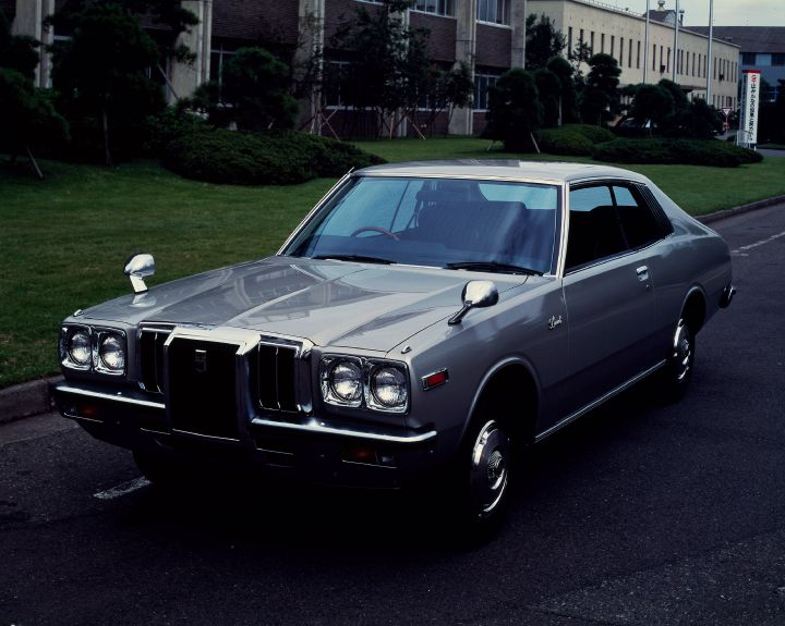 A vintage gray sedan is parked on a street near a grassy area. The car features classic design elements, such as chrome trim and distinctive headlights, with a backdrop of industrial buildings.
