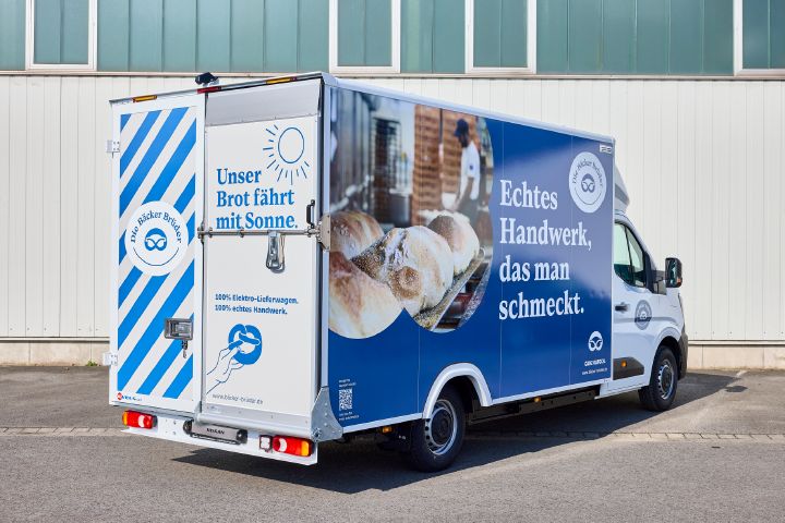 Blick von schräg hinten auf den Nissan Interstar-e mit Bäckereikoffer, die blau-weiße Beklebung zeigt die Schriftzüge "Unser Brot fährt mit Sonne" und "Echtes Handwerk, das man schmeckt"