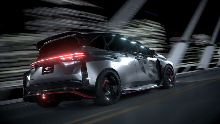 A silver and black two-tone sports car, the Aura Nismo RS Concept, seen from the rear as it speeds across a bridge. The vehicle is angled toward the right front, with bright red taillights illuminated. The rear features a black plate with white text reading 'AURA NISMO RS CONCEPT,' and a red accent line runs along the lower bumper. The background shows streaks of light, emphasizing motion and speed