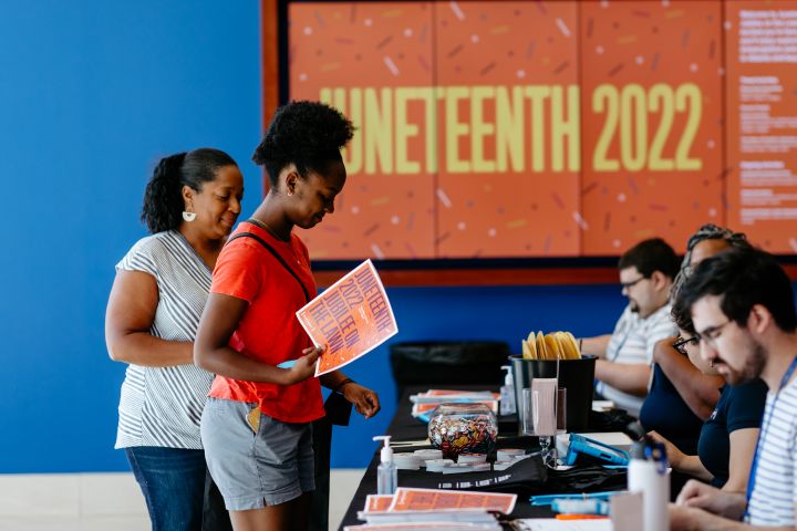 Two women, holding Juneteenth event flyers, stand Infront of a Juneteenth booth with volunteers sitting behind.