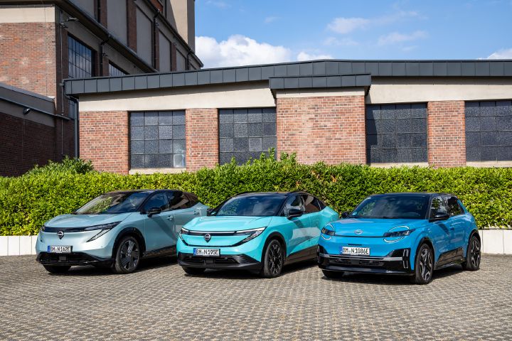 Vor einem Backsteingebäude und einer grünen Hecke stehen nebeneinander die drei Nissan EV-Modelle Nissan Ariya, Nissan Leaf und Nissan Micra, 3/4 Front