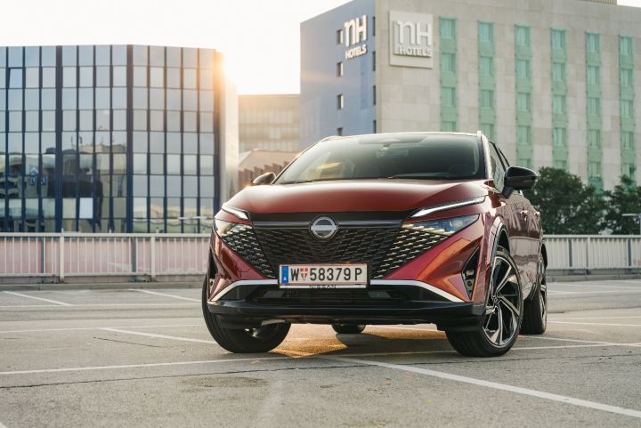 Frontaler Blick von unten auf einen Nissan Qashqai in Fuji Sunset Red auf einem Parkplatz vor einem Hotelgebäude