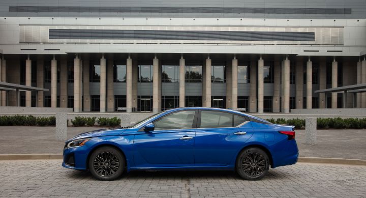 Driver-side profile shot of the Rear driver-side 3/4 shot of the 2024 Altima SV Special Edition driving down a wooded road in front of a building with columns