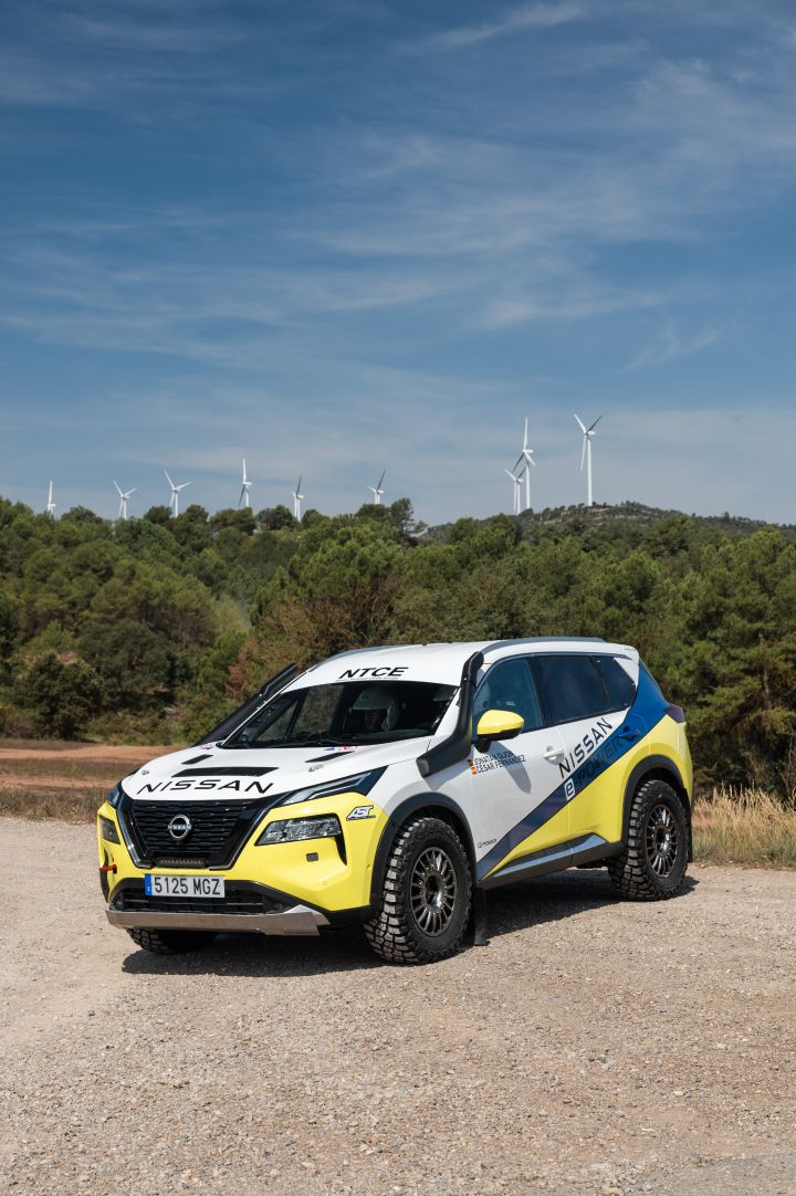 Standaufnahme von schräg vorne des Nissan X‑Trail Rennwagens für die Baja Aragon Rallye 2024 auf Schotteruntergrund und vor einem bewaldeten Hügel