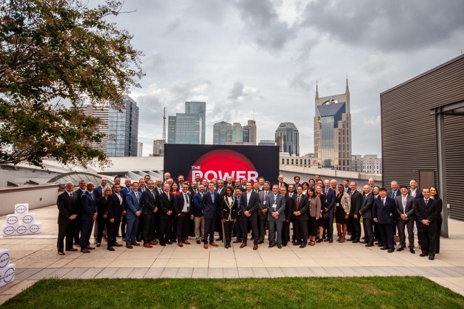 A group of people in business dress stand in front of a billboard on the roof of a building overlooking a city.