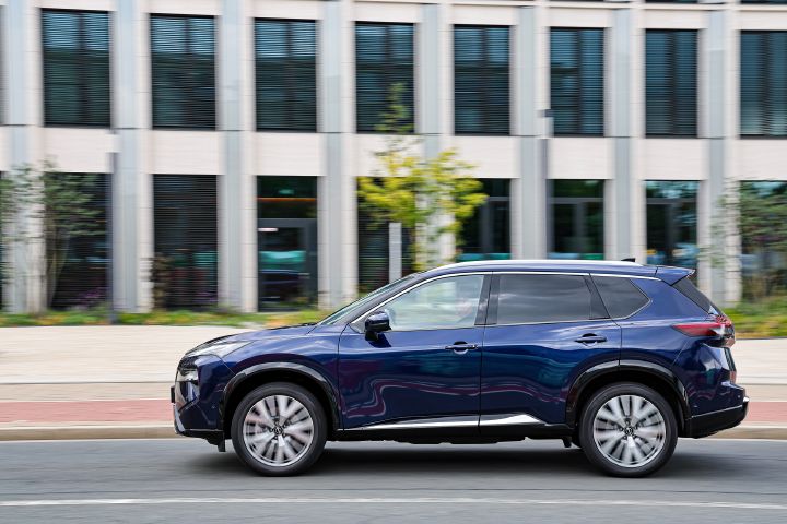 Vor einem hellen, modernen Gebäude mit vielen dunklen Fenstern fährt seitlich ein dunkelblauer Nissan X‑Trail vorbei