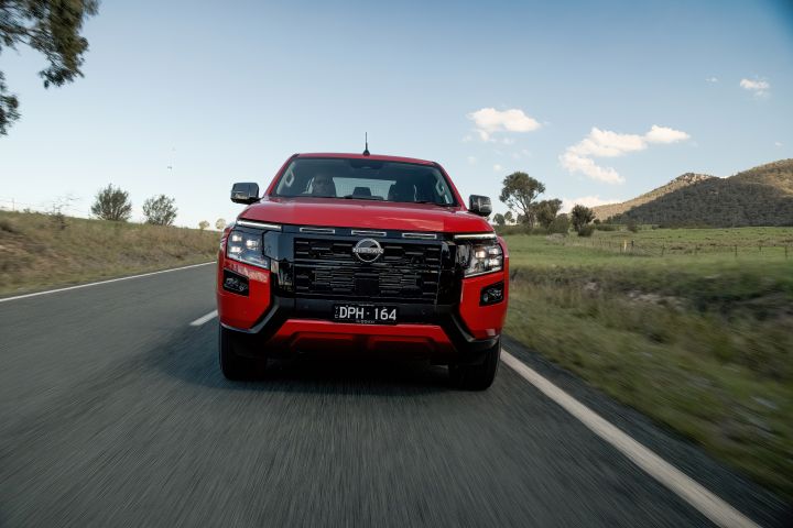 Underbody close-up of a 2026 Nissan Navara ST‑X dual cab finished in Outback Red, highlighting suspension components and rear tyre articulation on uneven dirt terrain.