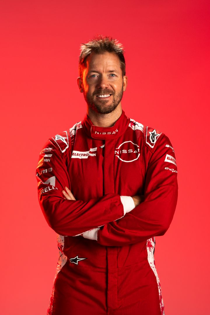 Sam Bird lächelt in seinem roten Rennanzug des Nissan Formula E Teams mit vor der Brust verschränkten Armen vor einem roten Hintergrund.