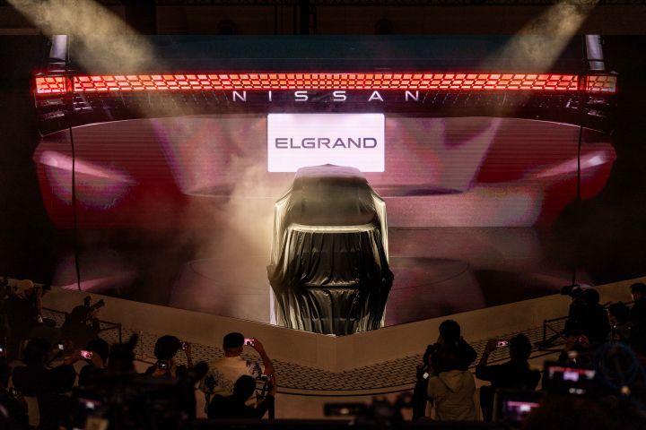 At the center of the darkened stage, the all-new Elgrand is positioned, covered with a black cloth, while the background screen displays the rear view of the Elgrand. In the foreground, members of the press are gathered, taking photos with their cameras
