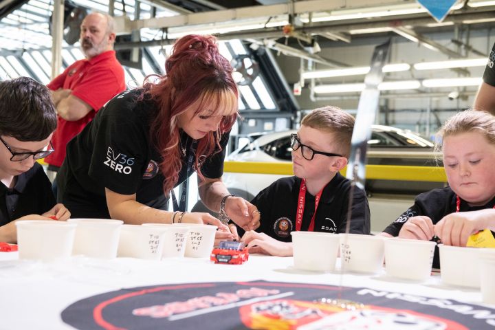 A Nissan employee assists school children in building a LEGO Nissan Qashqai, with the real production line visible in the background.