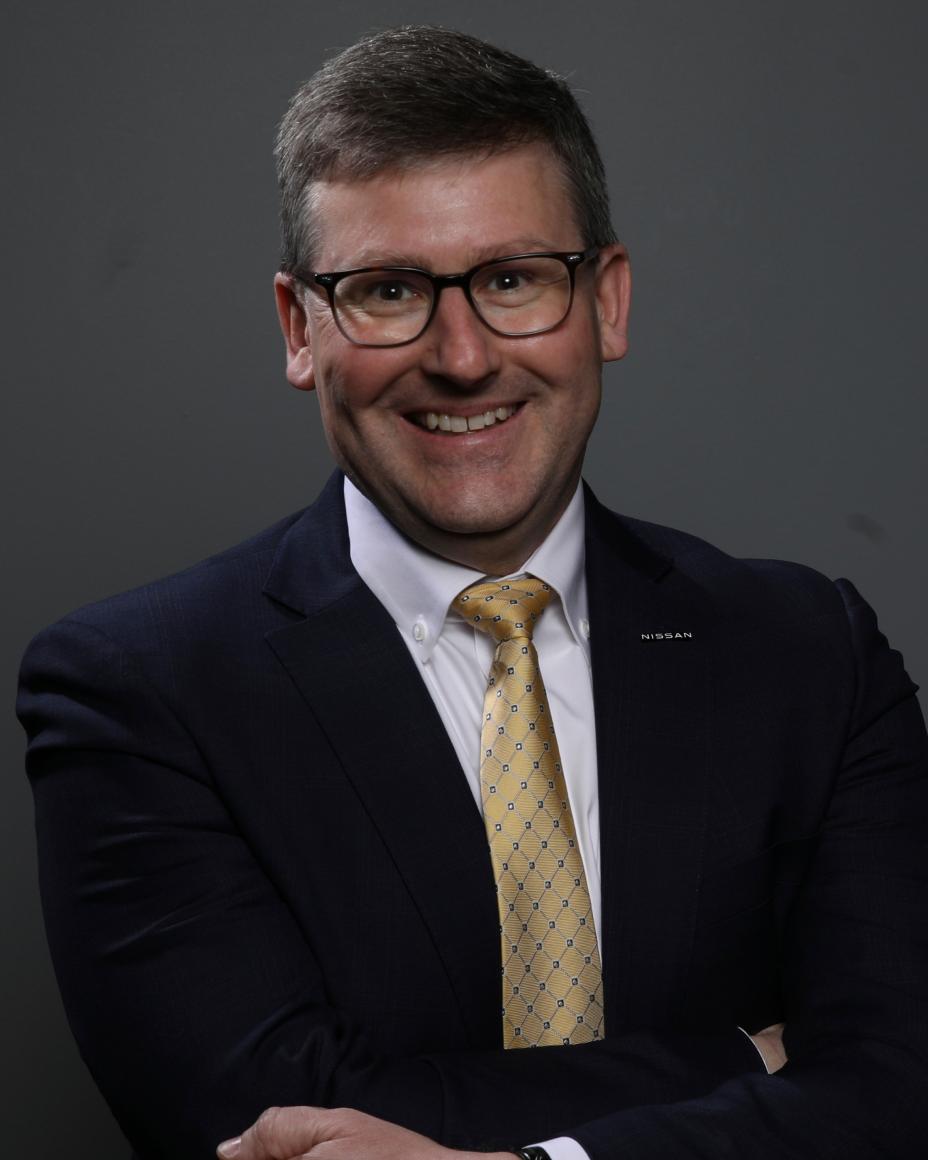 John, smiling from the chest up, in a black suit, against a gray background.