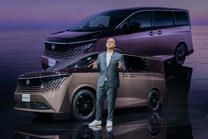 On the stage, the all-new Elgrand in Fuji Dawn and Shigoku two-tone colors is positioned diagonally, facing slightly to the left. In front of the car stands CEO Ivan Espinosa in a suit, gesturing with both hands while delivering a presentation. The background displays a large visual of the same vehicle.