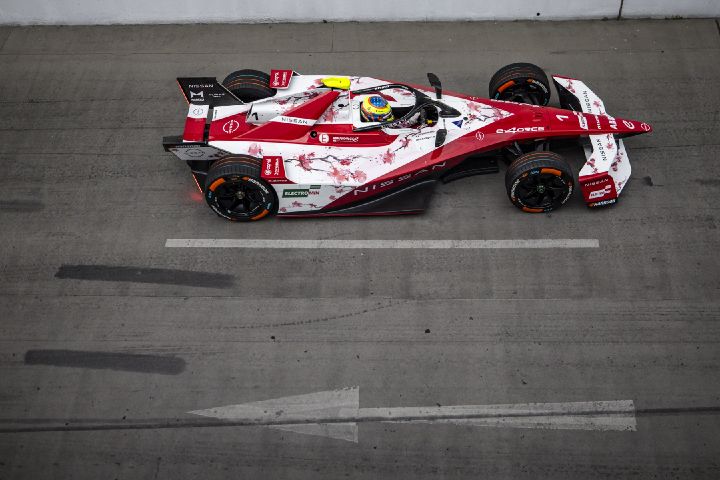 Eine Draufsicht auf einen rot-weißen Nissan Formel-E-Rennwagen, der auf einer Betonfahrbahn nach rechts fährt. Im Cockpit ist Oliver Rowland zu sehen, im Hintergrund eine weiße Wand und Fahrbahnmarkierungen.