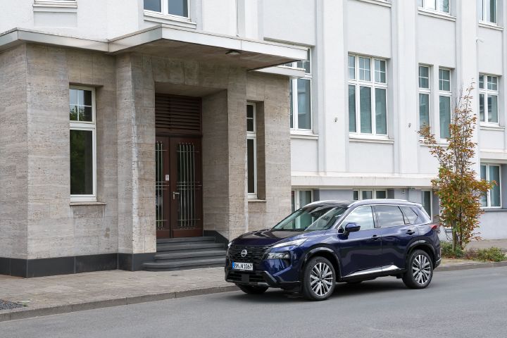 Ein dunkelblauer Nissan parkt am Straßenrand vor einem großen weißen Gebäude mit hellbraun abgesetztem Eingangsbereich