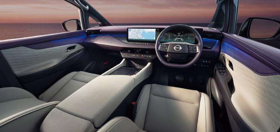Interior view of the All-new ELGRAND front cabin, photographed from behind the front seats. The cabin features a premium two-tone ivory and dark color scheme with high-quality materials extending across the dashboard and door panels. A wide digital display spans the driver area, showing navigation information and a digital instrument cluster. The large center console extends between the front seats and incorporates a broad armrest and storage area. Blue ambient lighting runs along the dashboard and door trims, creating a modern and sophisticated atmosphere. Through the windshield, a sunset seascape is visible, enhancing the sense of openness and refinement within the cabin.