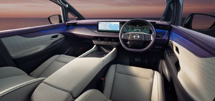 Interior view of the All-new ELGRAND front cabin, photographed from behind the front seats. The cabin features a premium two-tone ivory and dark color scheme with high-quality materials extending across the dashboard and door panels. A wide digital display spans the driver area, showing navigation information and a digital instrument cluster. The large center console extends between the front seats and incorporates a broad armrest and storage area. Blue ambient lighting runs along the dashboard and door trims, creating a modern and sophisticated atmosphere. Through the windshield, a sunset seascape is visible, enhancing the sense of openness and refinement within the cabin.