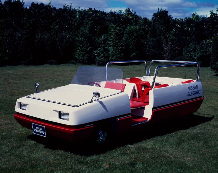 A vintage open-top Nissan electric vehicle with red and white color scheme is parked on a grassy field next to the edge of a forest.