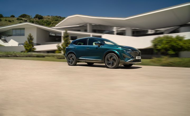 Ein dunkelgrün-blauer Nissan Qashqai mit e‑Power-Antrieb der nächsten Generation fährt bei Sonnenschein über eine Straße vor einem hellen Haus, der Hintergrund ist durch die Geschwindigkeit leicht verschwommen