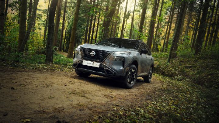 Modderige Ceremic Grey Nissan X-Trial N-Trek op een helling in een donkergroen bos met hoge bomen in de achtergrond