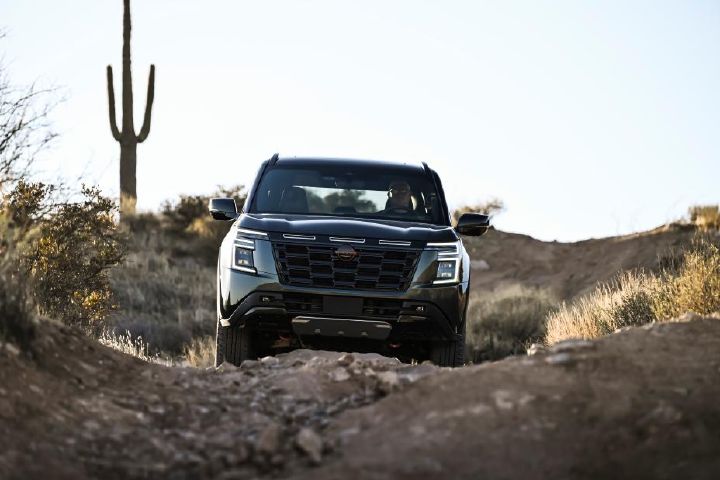 Front view of a 2025 Nissan Armada PRO-4X driving on uneven, rocky terrain during sunset, a cactus is behind.