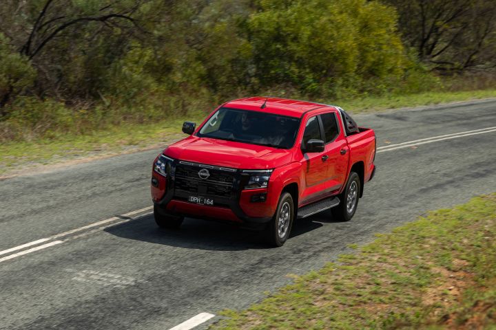 High-angle front three-quarter view of a 2026 Nissan Navara ST-X dual cab in Outback Red, driving on a sealed two-lane road bordered by grass and dense roadside vegetation.