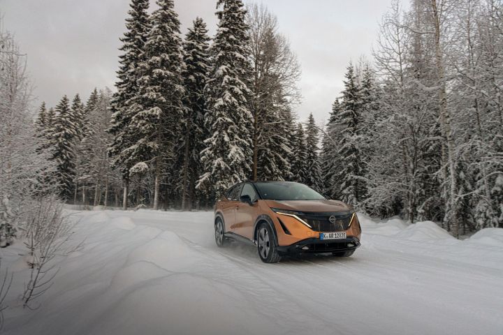 Elektroauto Nissan Ariya e‑4ORCE in Akatsuki Copper mit Dach und Außenspiegel in Pearl Black fährt auf schneebedeckter Straße mit verschneitem Wald im Hintergrund, 3/4 Front