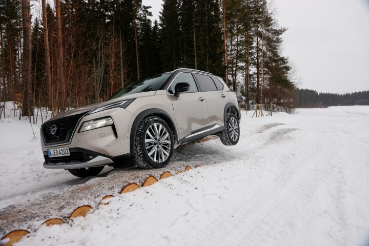 Frontansicht eines silbernen Nissan X‑Trail e‑Power e‑4ORCE, der über schneebedeckte Baumstämme fährt, ein Rad schwebt frei in der Luft