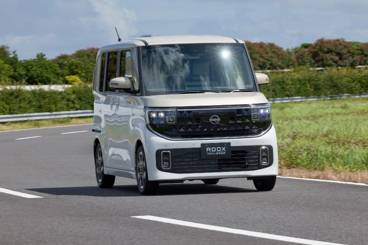 The new Nissan Roox kei car, in white, is driving forward on a paved road. The vehicle features a boxy design with large windows, and the background includes greenery such as trees and bushes under a clear blue sky.