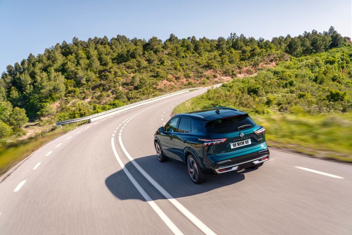 Heckansicht eines dunkelgrün-blauen Nissan Qashqai mit e‑Power-Antrieb der nächsten Generation, der an einem sonnigen Tag um eine Rechtskurve fährt mit grünen Bäumen und Büschen auf beiden Seiten der Straße