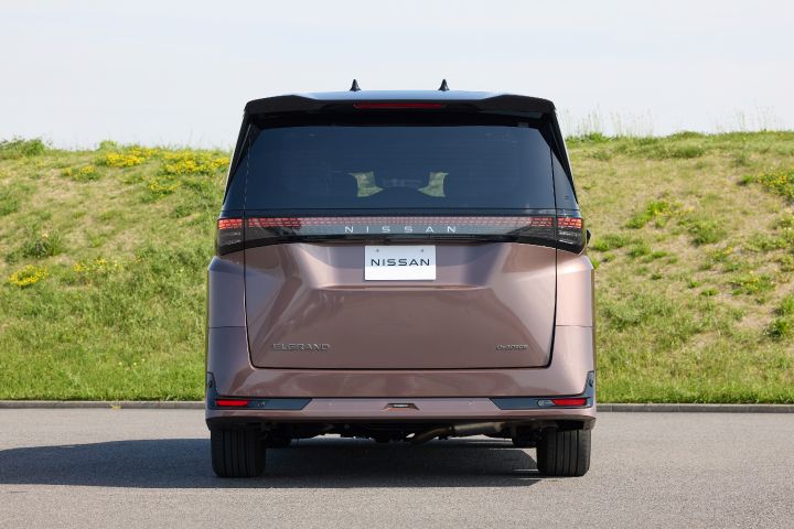 新型エルグランドが、正面リアビューで静止している。車両は中央に対称的に配置され、ブラックルーフと一体化したリアウインドウ、横一文字に広がるLEDテールランプ、中央の「NISSAN」ロゴが強調されている。背景には緑の草地と黄色い花が点在する土手が広がり、上空には淡い青空が広がる明るい屋外環境で、手前には灰色の舗装路面が見える。