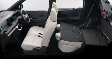 「日産サクラ」の車内を、後席側から前方に向けて撮影した画像。明るいグレーの前席と後席シートが配置され、前方には黒を基調としたインパネと右側配置のハンドルが見える。背景はスタジオの白背景で、室内空間全体が確認できる。