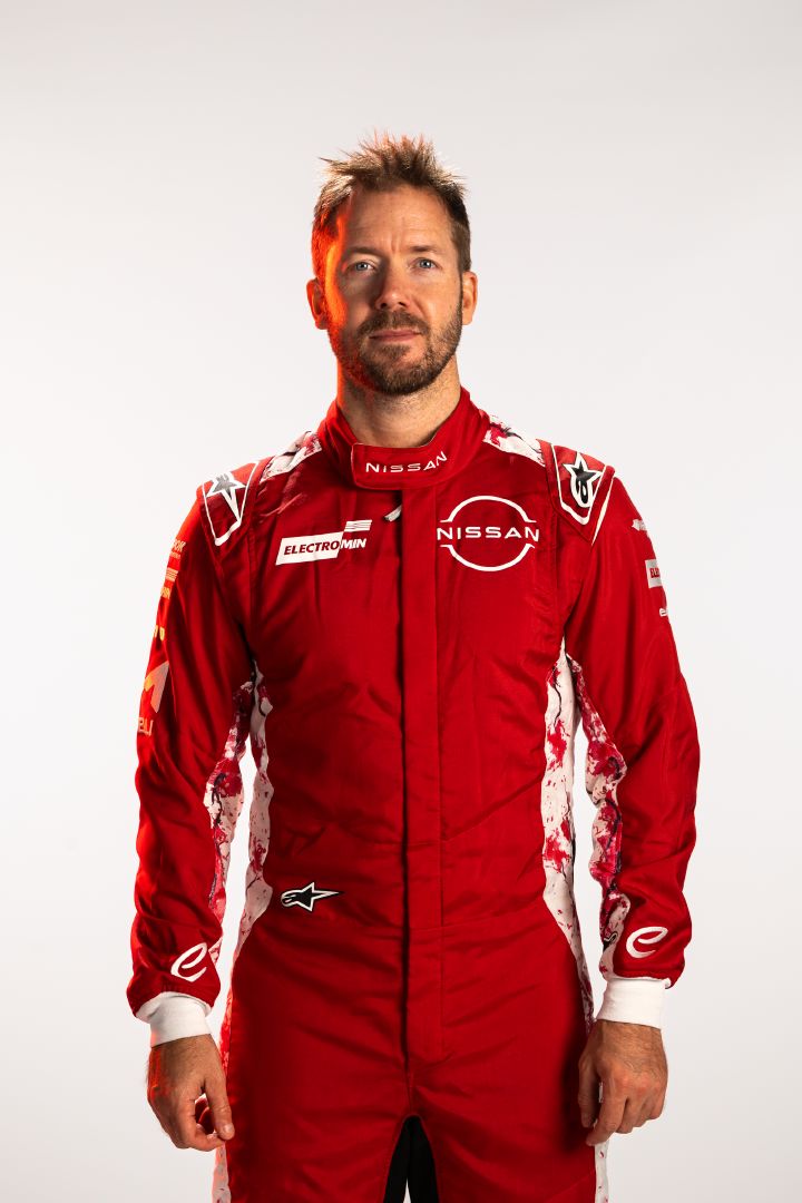 Sam Bird im roten Rennanzug des Nissan Formula E Teams mit den Armen locker an den Seiten herabhängend vor einem weißen Hintergrund.
