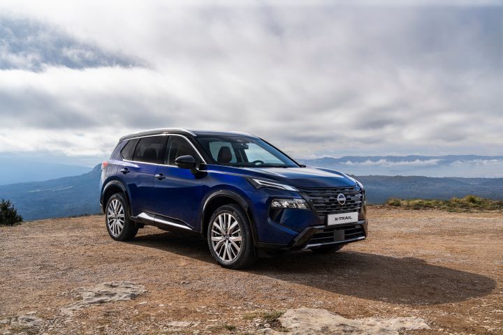 Neuer 2026 Nissan X‑Trail in der Karosseriefarbe Universal Blue, geparkt auf einem felsigen, erhöhten Gelände mit bewölktem Himmel und Bergen in der Ferne im Hintergrund. Das Fahrzeug verfügt über schlanke Scheinwerfer, Chromakzente und große Leichtmetallfelgen, die sein modernes Design und seine Geländegängigkeit unterstreichen.
