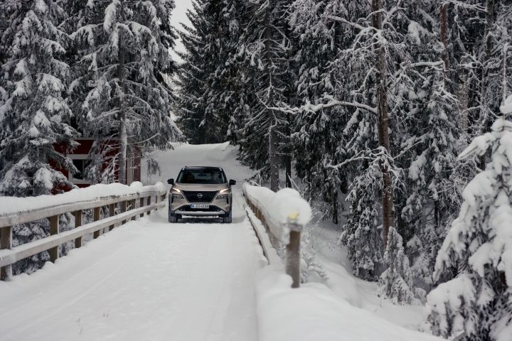 Silberner Nissan X‑Trail e‑Power e‑4ORcE fährt auf schneebedeckter Brücke mit verschneitem Wald und Holzhütte im Hintergrund, Front