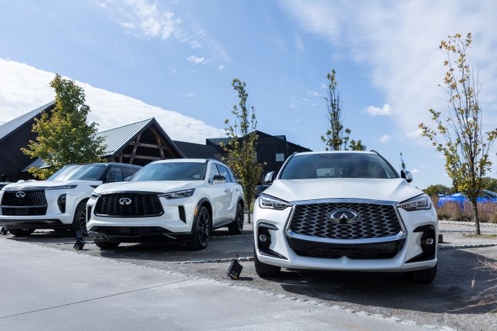 Three white INFINITI cars parked at an angle in front of a large venue building, the sky is blue with streaks of clouds.