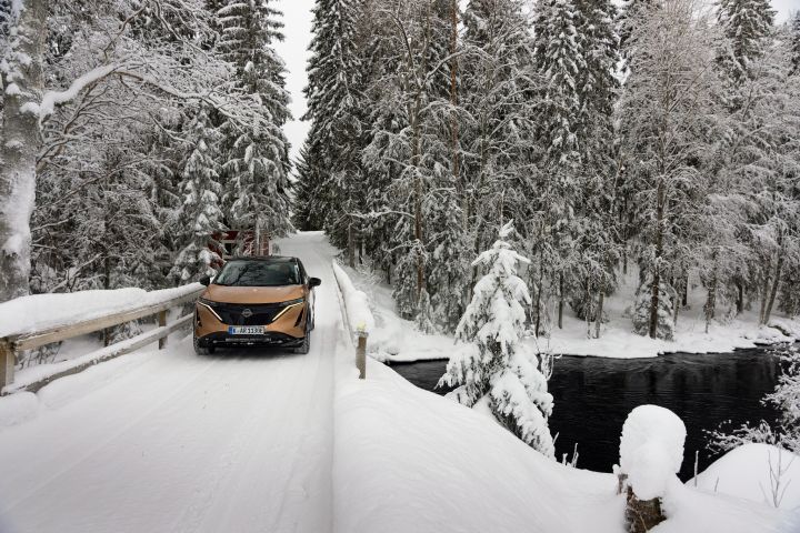 Elektroauto Nissan Ariya e‑4ORCE in Akatsuki Copper mit Dach und Außenspiegel in Pearl Black fährt auf schneebedeckter Brücke mit verschneitem Wald im Hintergrund, Front