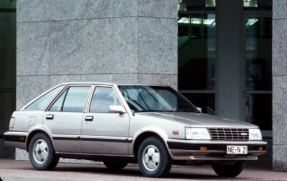 Ein silbergrauer Nissan Stanza in Front-/Seitenansicht vor einem großen Gebäude
