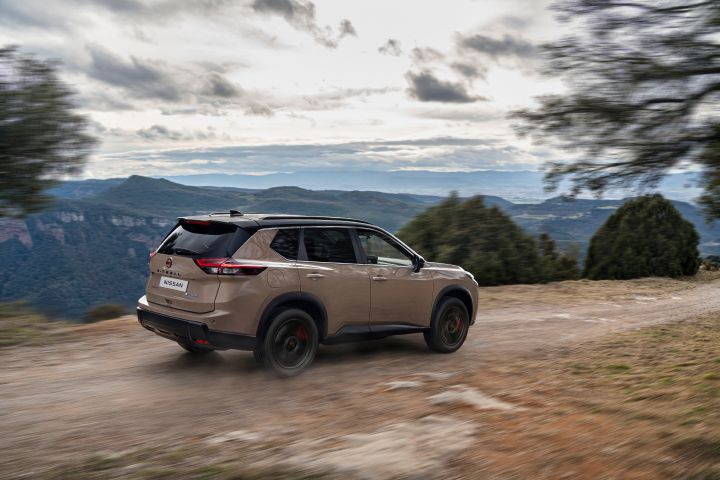 Der neue 2026 Nissan X‑Trail N-Trek in der Außenfarbe „Baja Storm” fährt auf einer unbefestigten Straße mit bergiger Landschaft und bewölktem Himmel im Hintergrund. Das Fahrzeug ist von hinten aufgenommen, wodurch sein schlankes Design und seine Geländegängigkeit besonders gut zur Geltung kommen.
