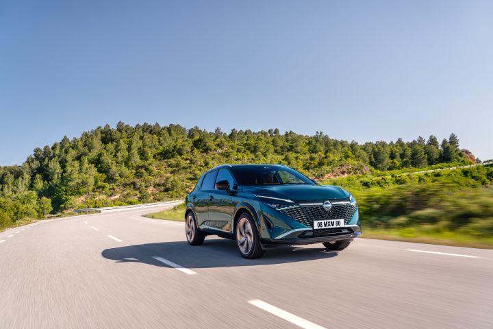 Fahraufnahme eines dunkelgrün-blauen Nissan Qashqai mit e‑Power-Antrieb der nächsten Generation auf einer sonnenbeschienenen Straße mit blauem Himmel und grünen Bäumen im Hintergrund