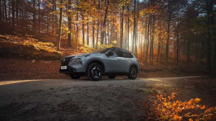 3/4 Frontansicht auf einen Nissan X‑Trail N-Trek auf einer Schlammstraße in einem herbstlichen Wald, die Sonne kommt durch die Bäume