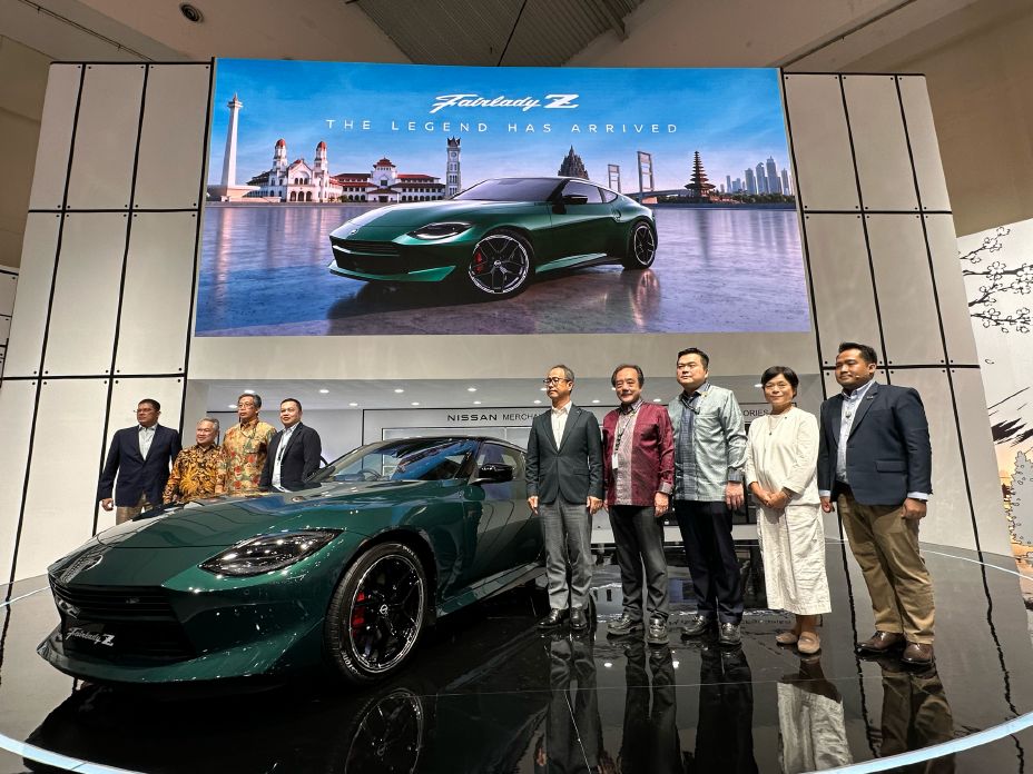 Group photo beside a Nissan vehicle displayed on a circular stage at an automotive exhibition, with a large digital screen showing the vehicle in the background.