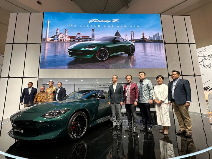 Group photo beside a Nissan vehicle displayed on a circular stage at an automotive exhibition, with a large digital screen showing the vehicle in the background.