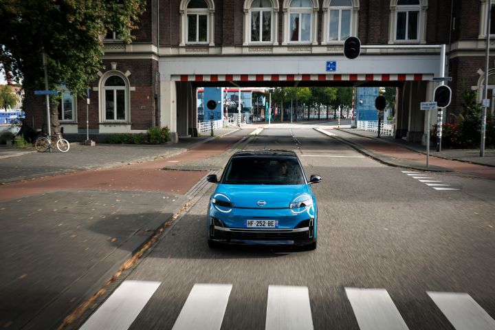 Frontale Fahraufnahme eines blauen Nissan Micra, der zwischen Gebäuden auf einen Fußgängerüberweg zufährt