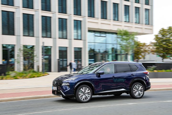 Ein dunkelblauer Nissan X‑Trail fährt auf einer Straße vor einem modernen Bürogebäude, neben dessen Eingang drei Menschen in Businesskleidung stehen