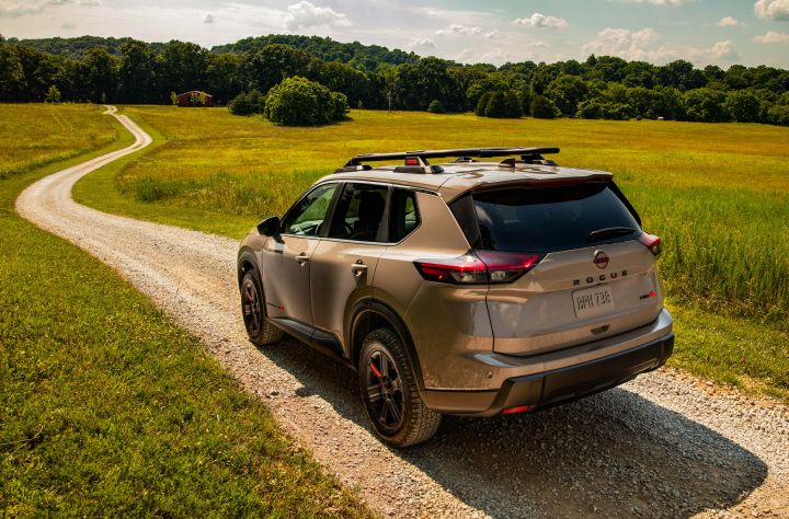 Left rear 3/4 view of a 2025 Nissan Rogue rock creek parked on a dirt road the winds down open fields and clustered trees.