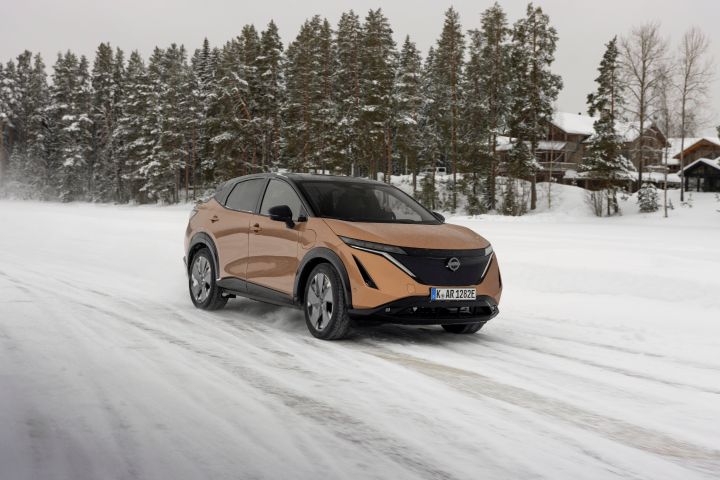 Elektroauto Nissan Ariya e‑4ORCE in Akatsuki Copper mit Dach und Außenspiegel in Pearl Black fährt auf schneebedeckter Straße mit verschneiten Bäumen und Holzhütten im Hintergrund, 3/4 Front