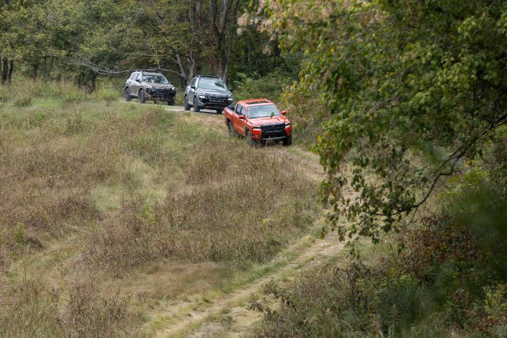 Two Nissan crossovers and a Nissan truck trail through an overgrown grass fields in the woods during the daytime.