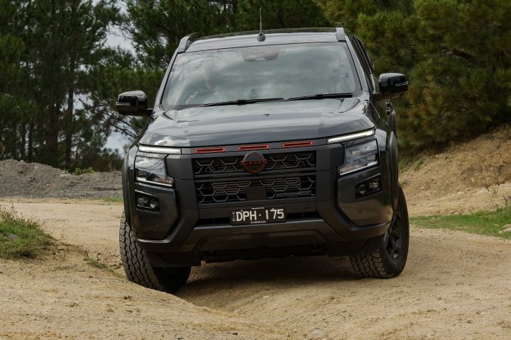 2026 Nissan Navara PRO-4X finished in Boulder Grey presented head‑on off‑road with red grille accents, visible lower protection and tyres loading into sandy ruts.