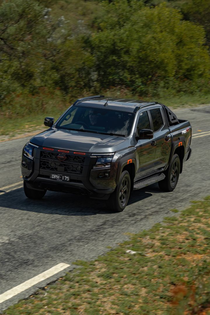 2026 Nissan Navara PRO-4X finished in Boulder Grey captured from a high front‑left angle on country bitumen with roof rails, side step and red grille tabs.